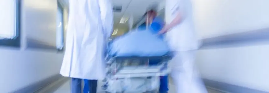 A motion blurred photograph of a patient on stretcher or gurney being pushed at speed through a hospital corridor by doctors & nurses to an emergency room