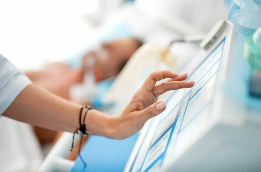 Close up of female hand with black bracelet touching display of breathing machine. Patient lying in hospital bed on blurred background