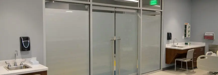large clean white hospital room with beds around the walls and a large glass door at the end of it
