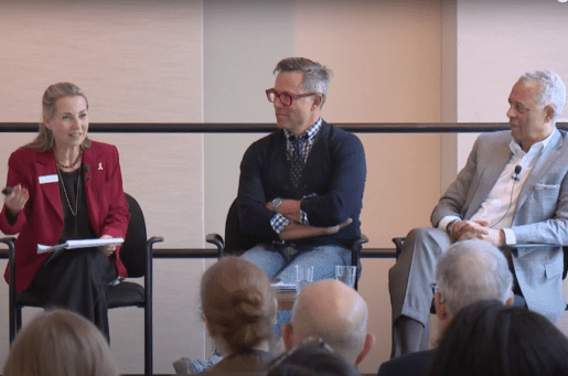 3 people sitting on a stage, a white woman in red, a white man in glasses, a black man in a gray suit