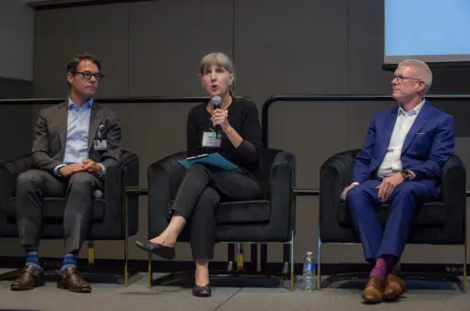 three older white people sitting on a stage, two men with woman speaking in middle