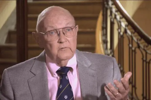 bald elder white man with wire aviator glasses, pink shirt and grey blazer