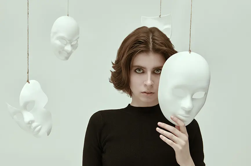 Theater of life. A girl in black clothes stands in a white room surrounded by various masks looking intently at the camera. Hypocrisy. Human roles. Mental disorders.