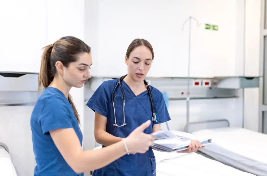 two young white women with brown hair and blue scrubs discussing chart
