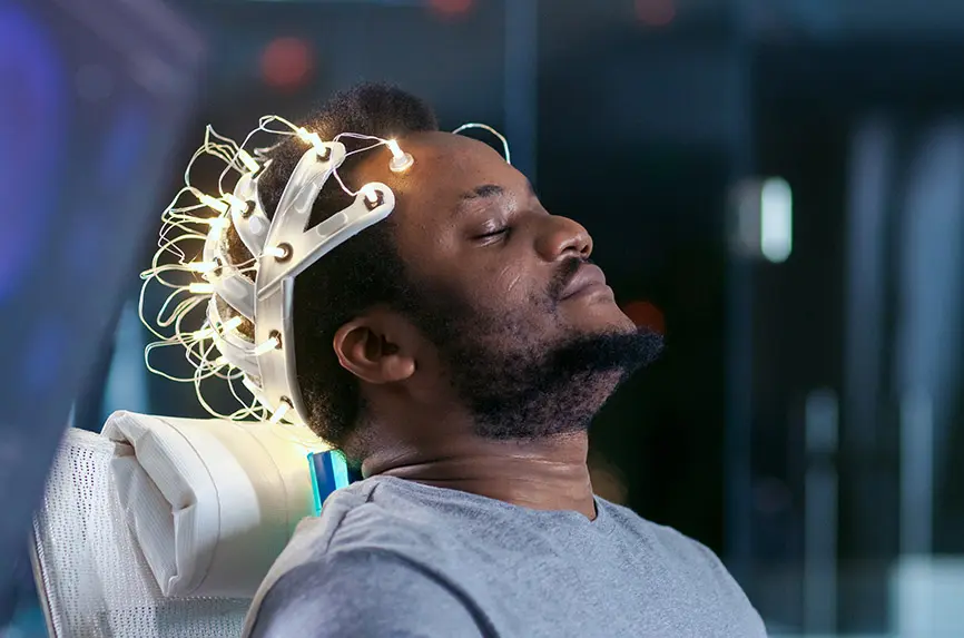 In Laboratory Man Wearing Brainwave Scanning Headset Sits in a Chair with Closed Eyes. Monitors Show EEG Reading and Graphical Brain Model. In the Modern Brain Study/ Neurological Research Laboratory.