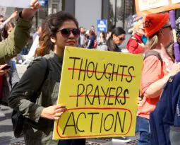 woman of color holding a yellow sign with the words "thoughts" and "prayers" crossed out, and the word "action" is circled in red marker