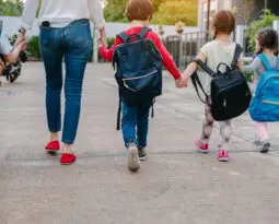 four young children wearing backpacks and walking hand in hand with a woman