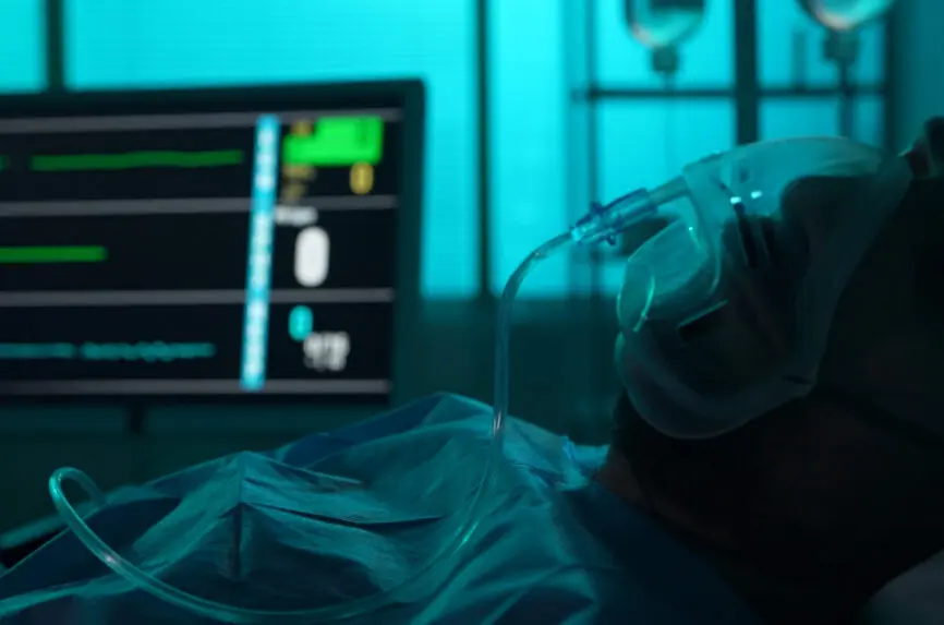 Close-up detail shot of a patient on breathing support dying in a hospital bed. His ICU monitor on the background displays loss of heart and brain activity.