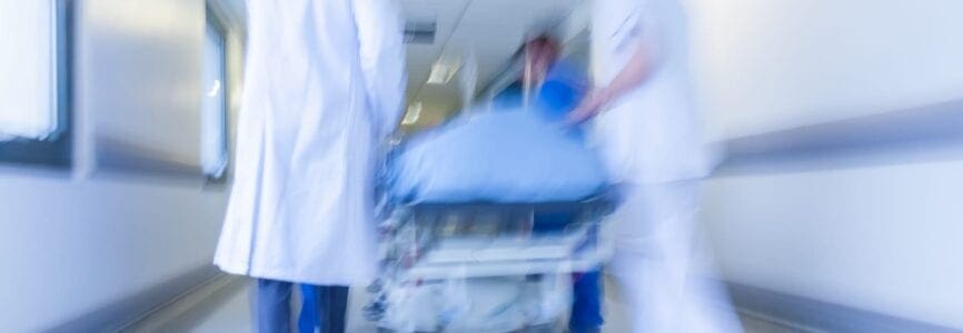 A motion blurred photograph of a patient on stretcher or gurney being pushed at speed through a hospital corridor by doctors & nurses to an emergency room