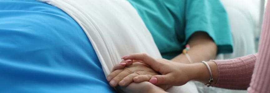 older woman laying in a bed with a younger woman holding her hand