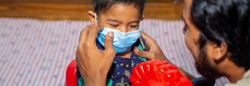 A young man is adjusting a baby's protective surgical face mask on the child's face for COVID 19 safety issue.