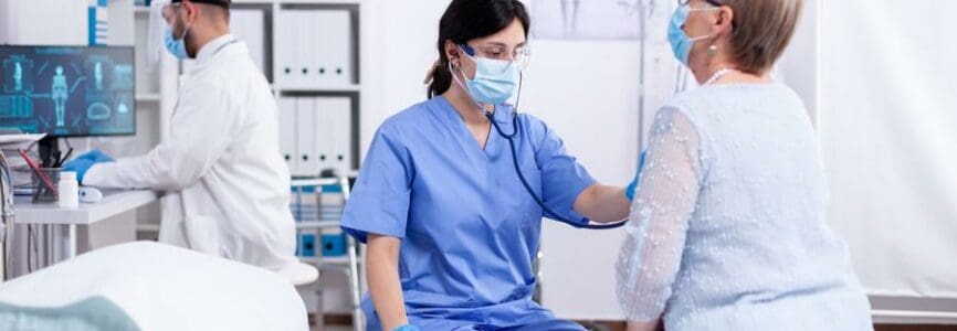 Cardiologist assistant listening heart of senior woman with stethoscope during consultation and wearing face mask against coronavirus outbreak. Medical examination for infections, disease.