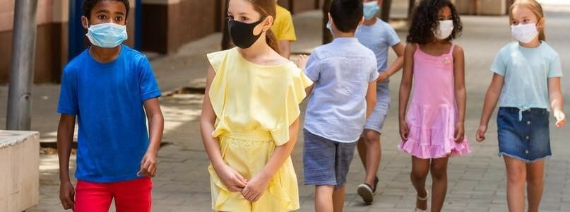 School children in protective medical masks walk along the street of a summer city