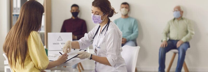 Mass vaccination of the population. Female doctor in a mask gives the patient documents to sign before giving her the Covid-19 antiviral vaccine. Background with people waiting in line for a vaccine.