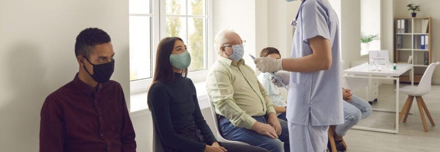 Young man doctor nurse in medical protective face mask talking to patients sitting and waiting in line before vaccination against covid-19 virus during pandemic in medical clinic office