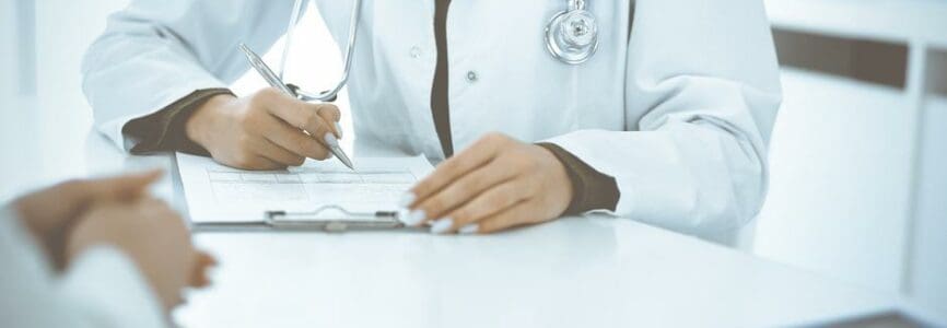 Unknown woman-doctor and female patient sitting and talking at medical examination in clinic, close-up. Therapist wearing green blouse is filling up medication history record. Medicine concept.