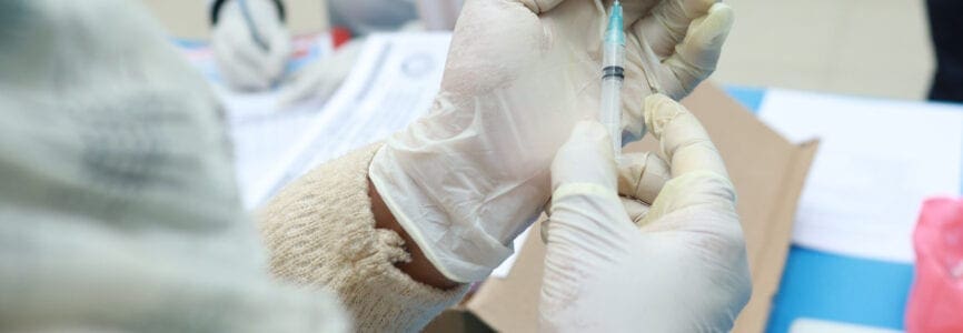 a medical doctor drawing vaccine from a vial in a syringe in a gloved hand with selective focus on vaccine.