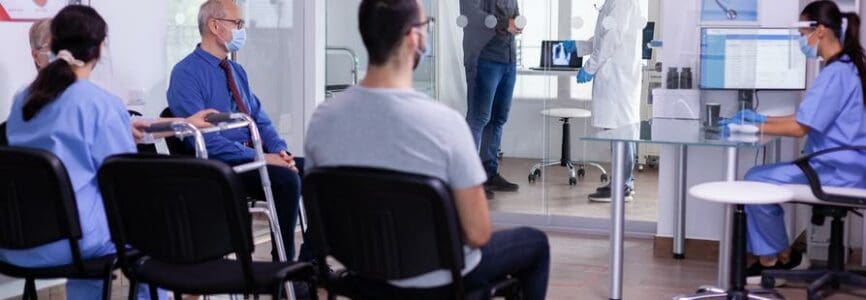 Crowded hospital waiting room with patients sitting on chairs respecting social distencing and nurse explaining treatment to elderly woman wearing face mask against covid-19. Doctor consultating man