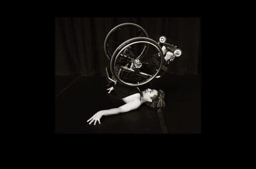 Black and white photo of wheelchair dancer Alice Shepard, with Alice, a white woman, on the ground and her wheelchair in the air.