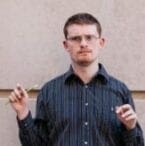 DJ Savarese, a white man with short, dark hair, poses contemplatively against a gray wall. His hands are raised before him as if in motion, one of them holding a pen.