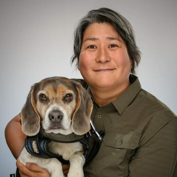 Karen Nakamura holds a beagle wearing a service dog vest in her arms. She has a relaxed smile on her face as both she and the dog gaze into the camera.