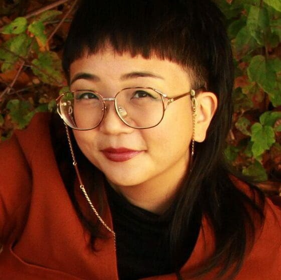 Jina, an Asian woman with short bangs, smiles at the camera against a background of ivy leaves. A thin gold chain hangs down from her glasses and her burnt orange cardigan matches her lipstick.