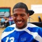 Lateef McLeod, a Black man wearing a blue jersey, is caught in the middle of laughing. His eyes are pressed shut and a smile stretches across his face.