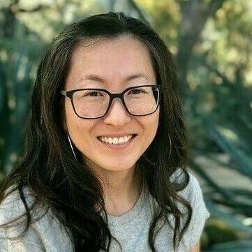 Mia, a korean woman wearing glasses and a gray shirt, sits in front of a garden full of succulent plants. She is smiling and her dark hair falls in waves over her shoulders.