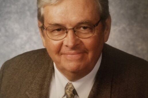 elder white man with rectangle wire frame glasses in a brown suit and grey background