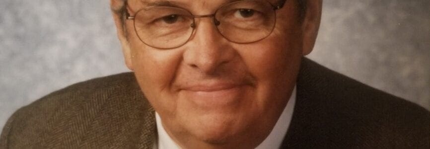 elder white man with rectangle wire frame glasses in a brown suit and grey background