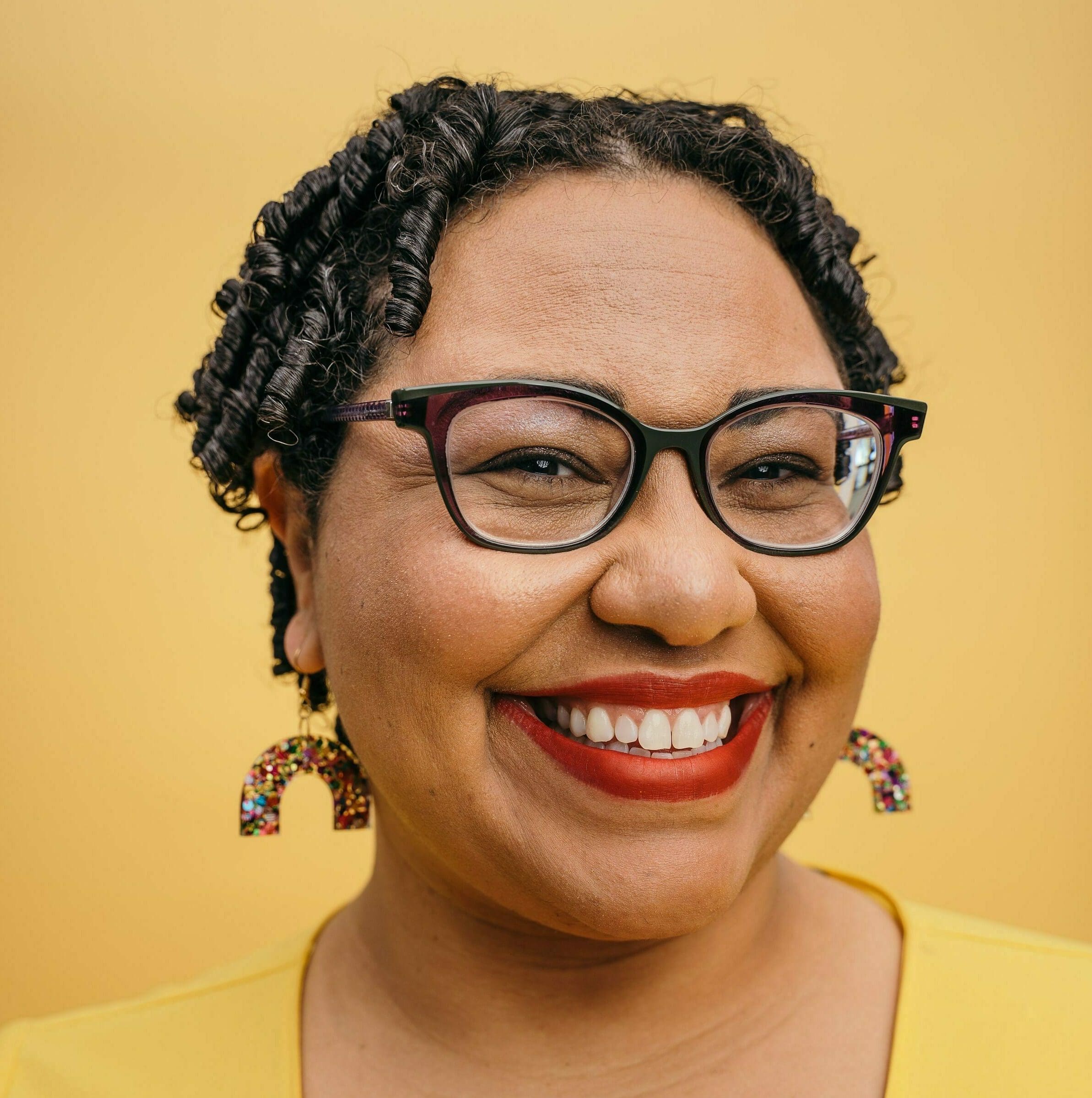 Fat Black light-skinned woman with glasses smiles at the camera. She has short black hair with defined curls, red lipstick, and multi-colored glitter earrings in a rainbow shaped. She wears a bright yellow top and the background is a mustard yellow.