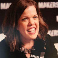 Leah, a white woman with wavy brown hair, smiles widely while talking into a microphone. She is wearing a black blazer and a chunky bronze necklace. 