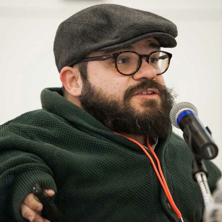 Joseph, a white man with glasses and a thick beard, speaks into a microphone. He is wearing a gray driving cap and seems to be talking animatedly.