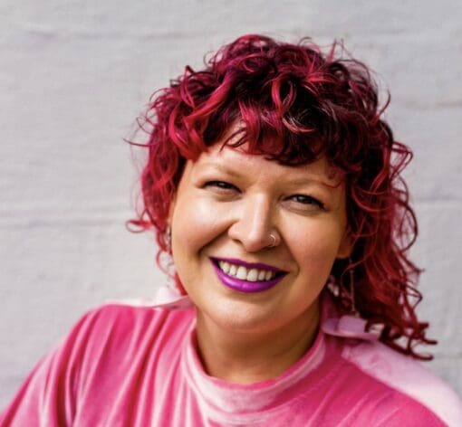 Jess, a white femme person with pink hair and pink lipstick smiles against a white wall. They are wearing a pink velvet garment.