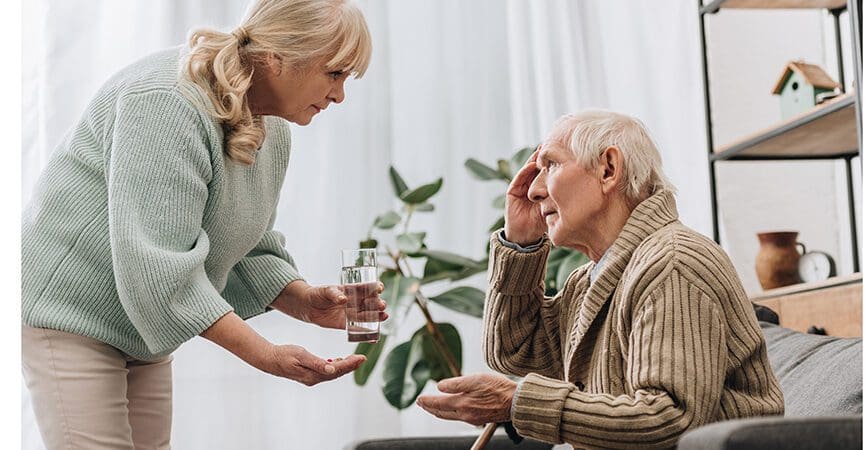 senior woman giving pills and glass of water to old man with walking stick