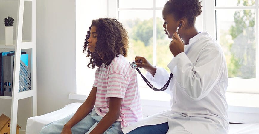 Female doctor listens to back of preteen girl patient to check her lungs, breathing and heartbeat. Doctor listens with stethoscope to African American teenage girl sitting on examination couch.