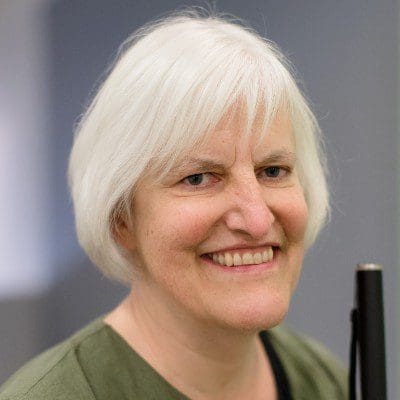 Georgina Kleege, a white woman with a short white bob haircut, beams at the camera with a wide smile, holding her white cane in front of her.