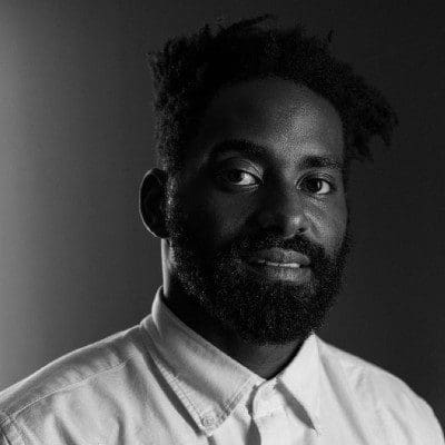 Black and white photo of Jerron Herman, a dark-skinned Black man with a beard, wearing a white button up looking into the camera with a slight smile. Photo by Fernando Villela