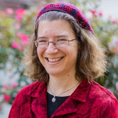 A portrait-style photo of Julia Watts Belser, a white, curly-haired woman, sitting happily in her wheelchair in front of a garden with pink flowers. She's wearing a patterned red blazer and red kippah (beret) to match.