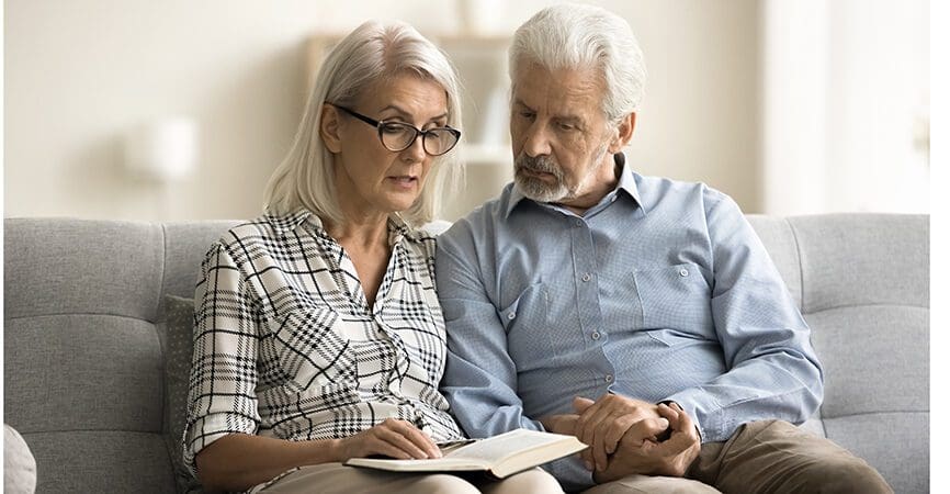 Caring senior wife reading book aloud to older husband, reviewing written notes in notebook. Elderly retired couple enjoying leisure, literature at home together, holding hands