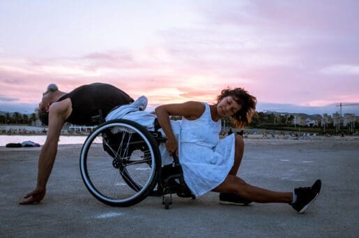 Two people dance under the sunset on a beach. On the left, a man leans back in his wheelchair with his arms dangling behind him, hands grazing the ground. In front of him, a woman grips the chair to press her back against his shins, stretching her own leg out in front of her.