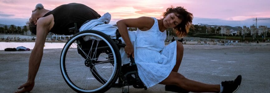 Two people dance under the sunset on a beach. On the left, a man leans back in his wheelchair with his arms dangling behind him, hands grazing the ground. In front of him, a woman grips the chair to press her back against his shins, stretching her own leg out in front of her.