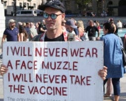 white man holding a white sign stating in red lettering "I will never wear a face muzzle, I will never take the vaccine"