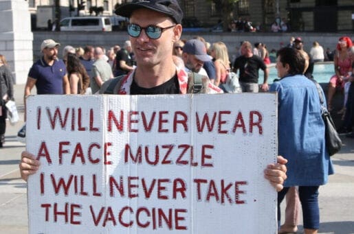 white man holding a white sign stating in red lettering "I will never wear a face muzzle, I will never take the vaccine"