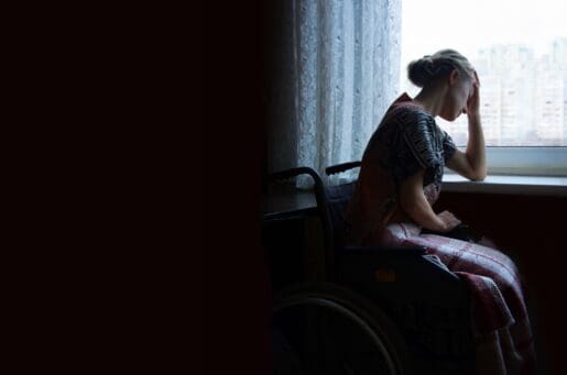 white woman with a bun sitting by window in wheelchair holding head in hand