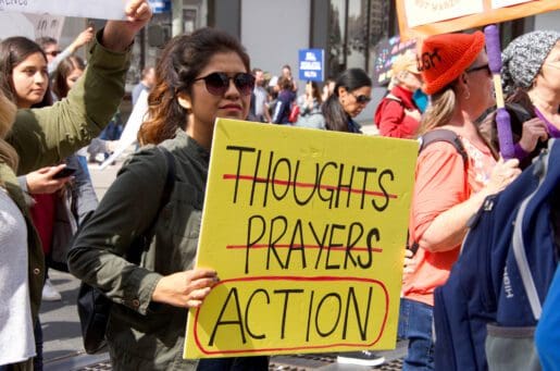 woman of color holding a yellow sign with the words "thoughts" and "prayers" crossed out, and the word "action" is circled in red marker