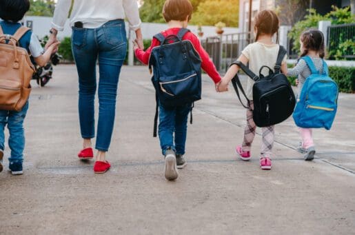 four young children wearing backpacks and walking hand in hand with a woman