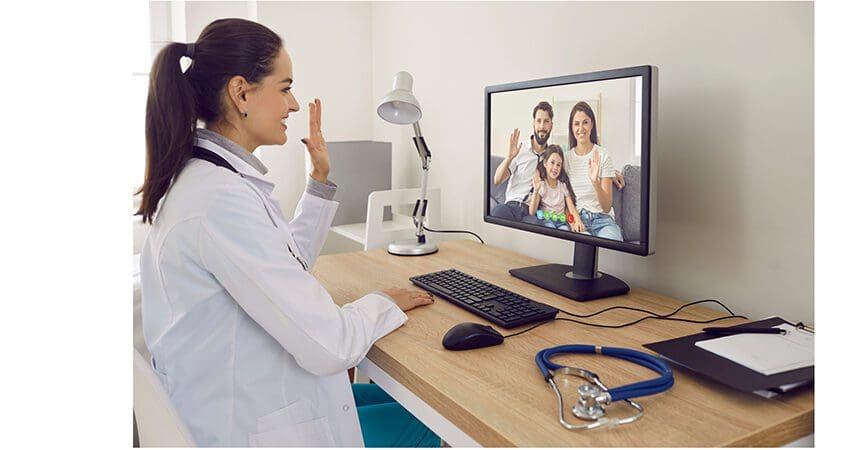 Online family doctor at desk with stethoscope smiling and waving hello at desktop computer screen greeting parents and child at telemedicine consultation via video call in modern convenient webcam app